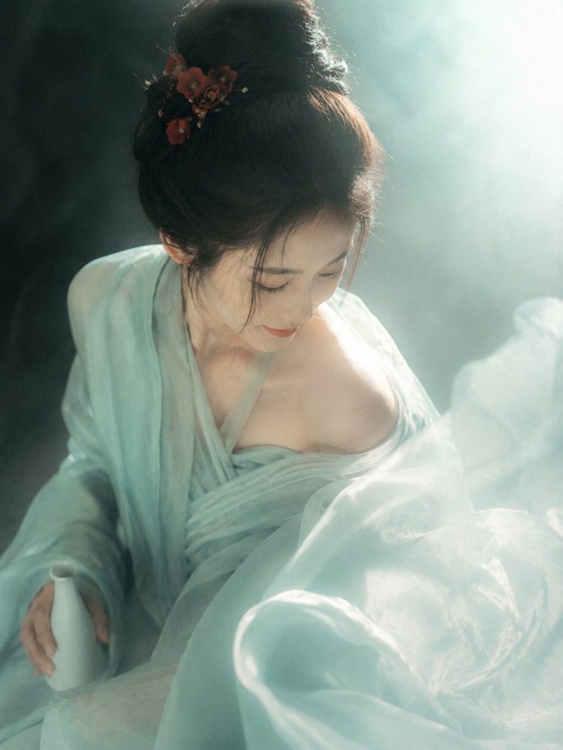 A beautiful woman in a flowing light blue dress with her hair in an updo, holding a small white vase.