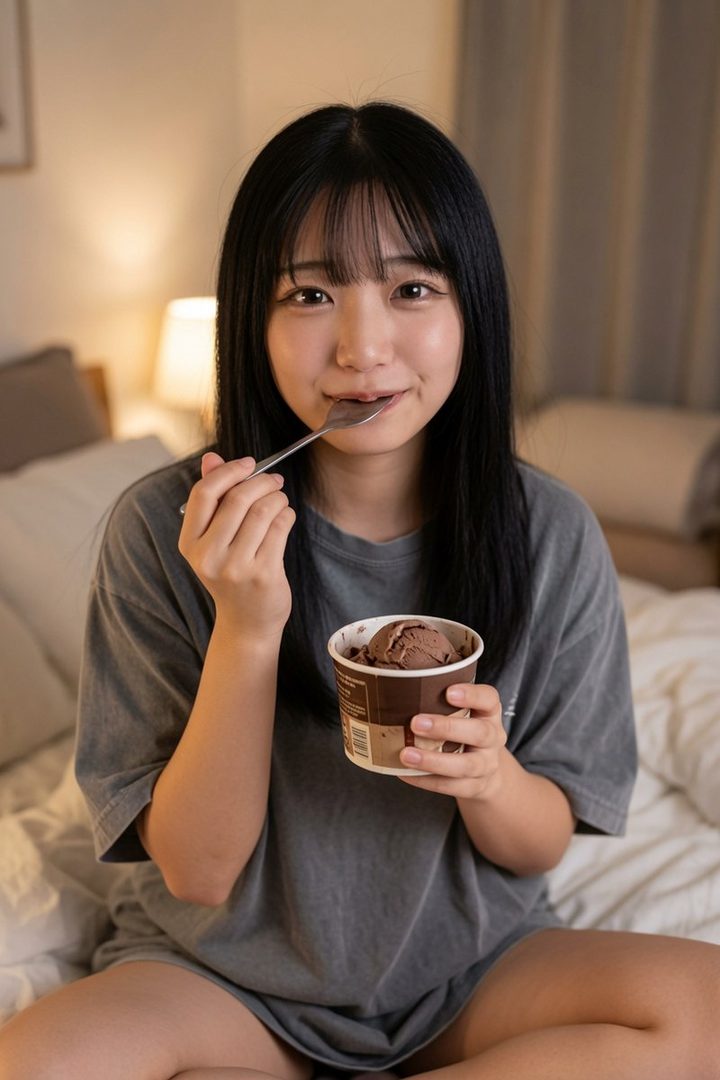 A young woman with long black hair and bangs is sitting indoors, eating chocolate ice cream from a cup with a spoon.
