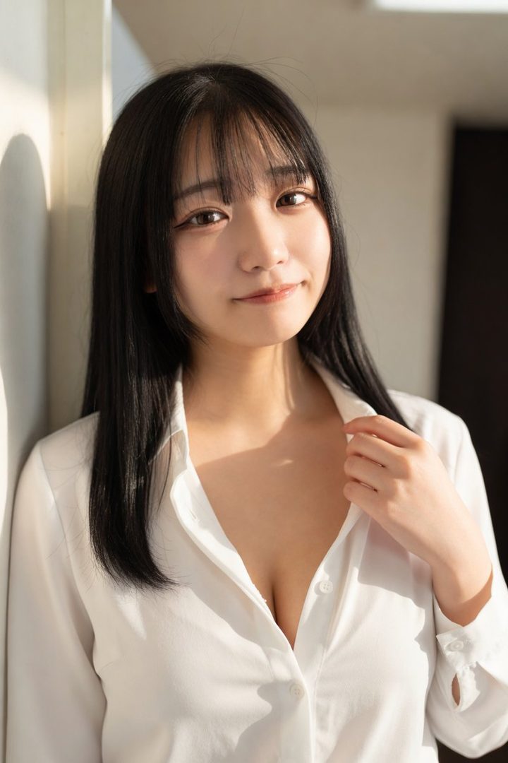 A young woman with long black hair and bangs wears a white blouse, looking at the camera with a gentle smile.