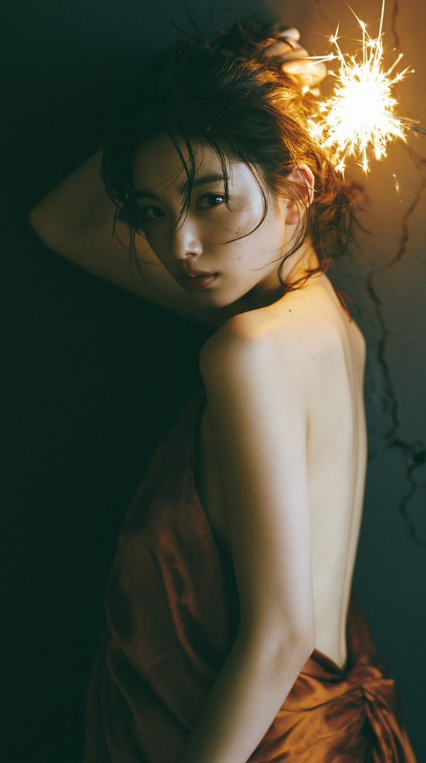 A woman with brown hair and a bare back holds a sparkler, looking dreamily at the camera.