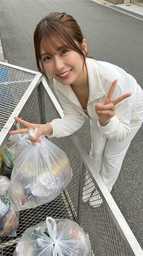 A cheerful woman in a white jumpsuit leans over a metal garbage bin, making peace signs with both hands, with clear plastic bags of trash visible.