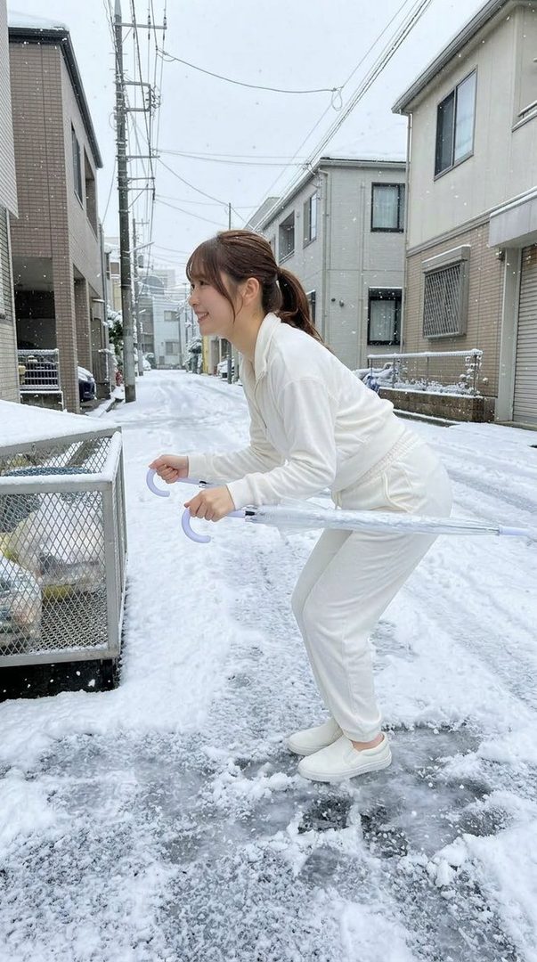 A young woman in a white tracksuit playfully holds an umbrella like ski poles on a snowy street.