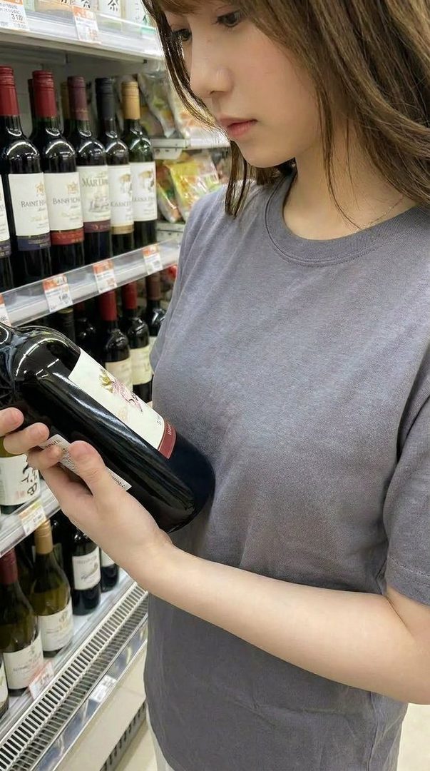 A young woman with brown hair in a grey t-shirt is looking at a bottle of red wine in a supermarket aisle.