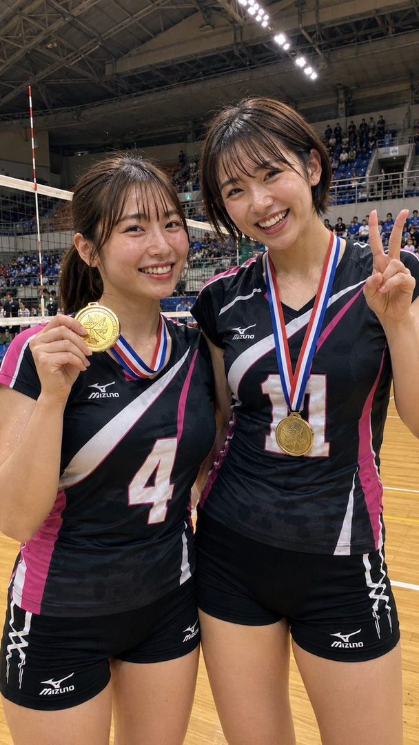 Two smiling women in black and pink volleyball uniforms proudly display their gold medals in an indoor sports arena.