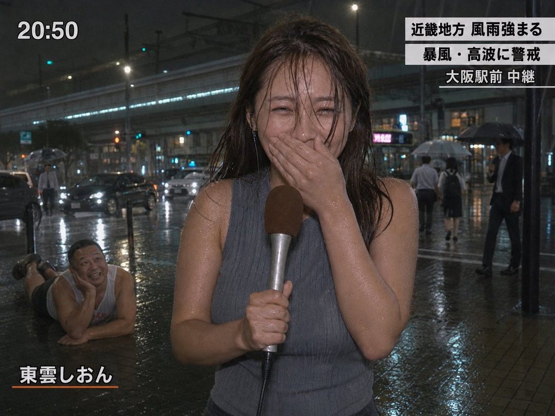 A woman reporter in a grey tank top laughs while holding a microphone in the rain at night, with a man lying on the wet ground next to her.
