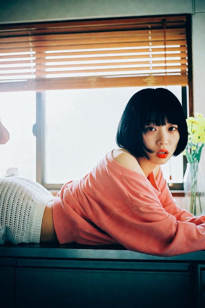 A young woman with black bob hair and red lipstick is lying on a counter in a pink off-shoulder sweater and white knit shorts, looking directly at the camera.