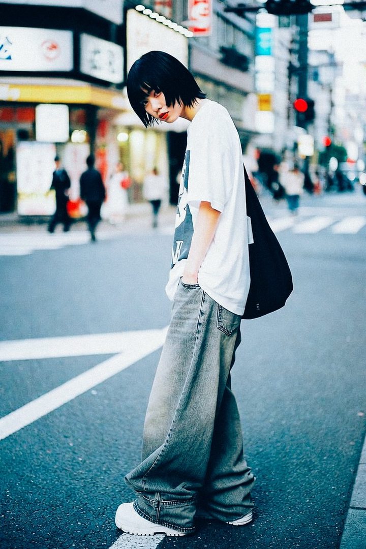 A person with short black hair and an oversized white t-shirt and baggy jeans stands on a city street, looking towards the camera.
