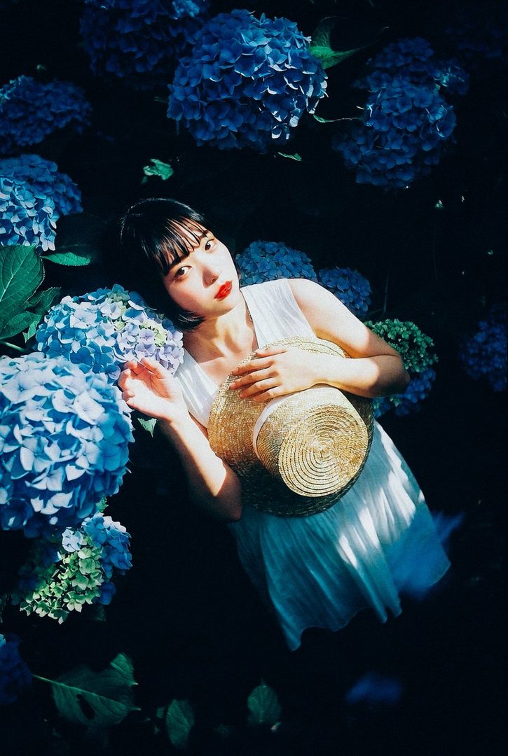 A woman with a bob haircut and a white dress lies among blue hydrangeas, holding a straw hat.