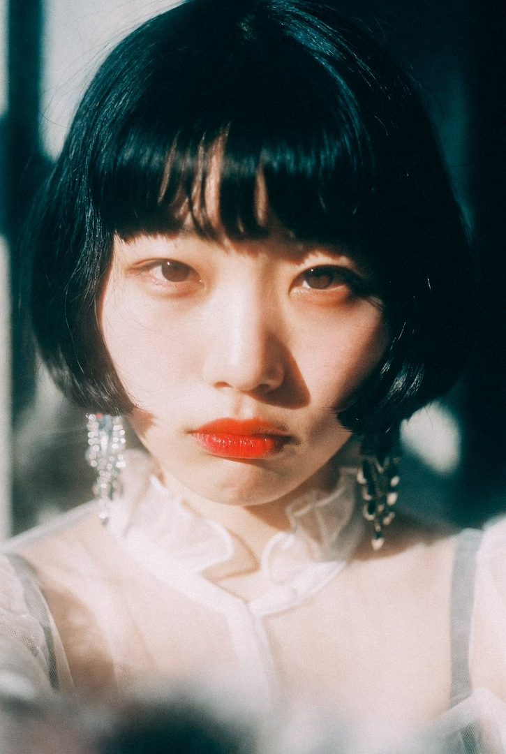 A close-up portrait of a woman with a black bob haircut and red lipstick, wearing a sheer top and large dangle earrings, with dramatic lighting.