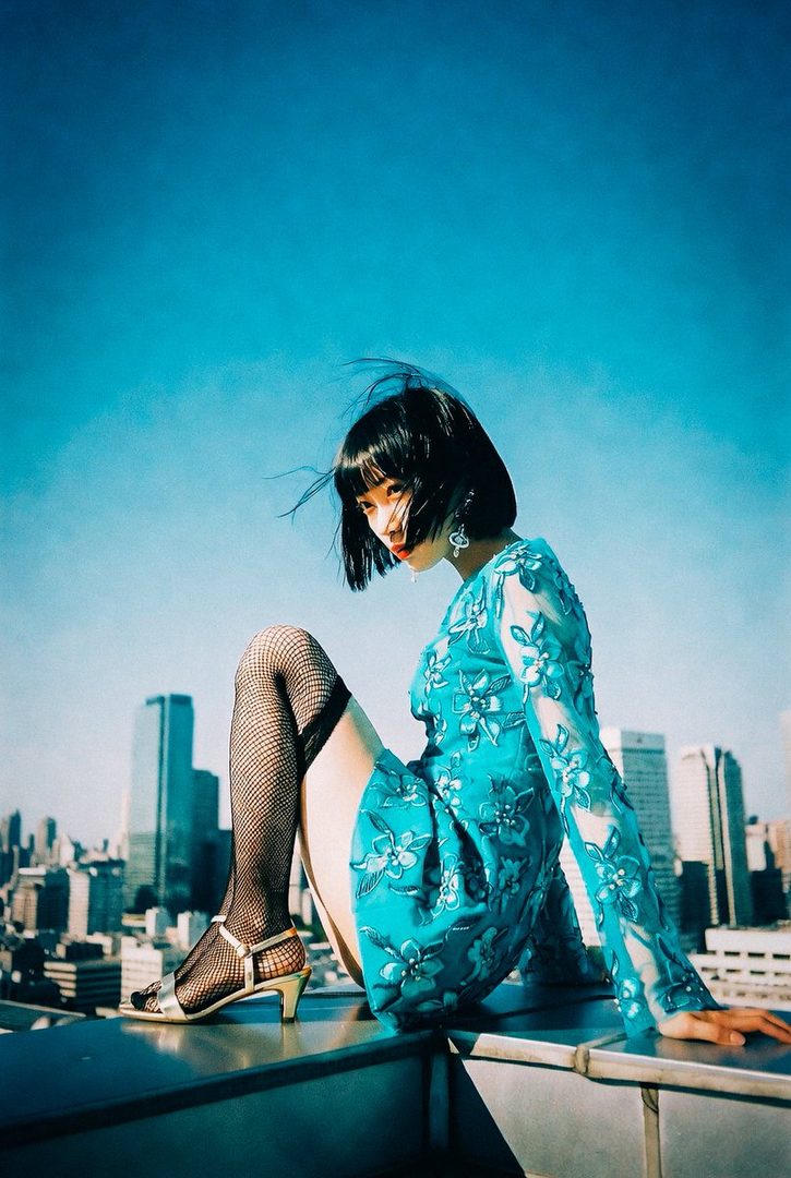 A woman with a bob haircut and a blue floral dress sits on a rooftop overlooking a city, with a bright blue sky in the background.