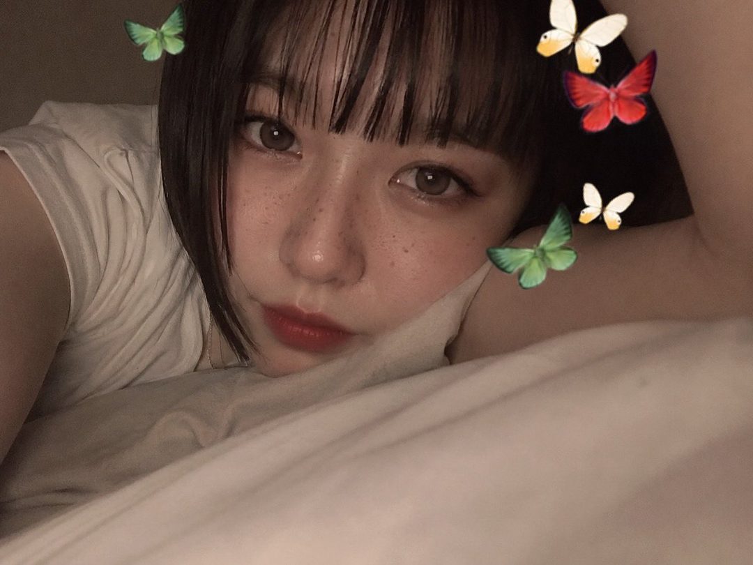 A close-up portrait of a woman with dark brown hair and bangs, lying down with freckles and red lipstick, surrounded by digital butterflies.