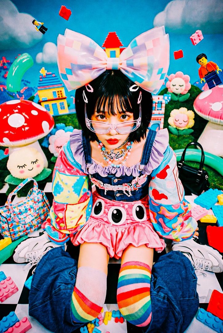A person with black bob hair and a large colorful bow wears a patchwork top, pink skirt with eyes, and rainbow socks, sitting in a whimsical, toy-filled setting.
