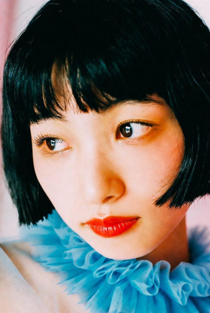 A close-up portrait of a woman with black bob hair and bangs, wearing a blue ruffled collar and red lipstick, looking thoughtfully to the side.