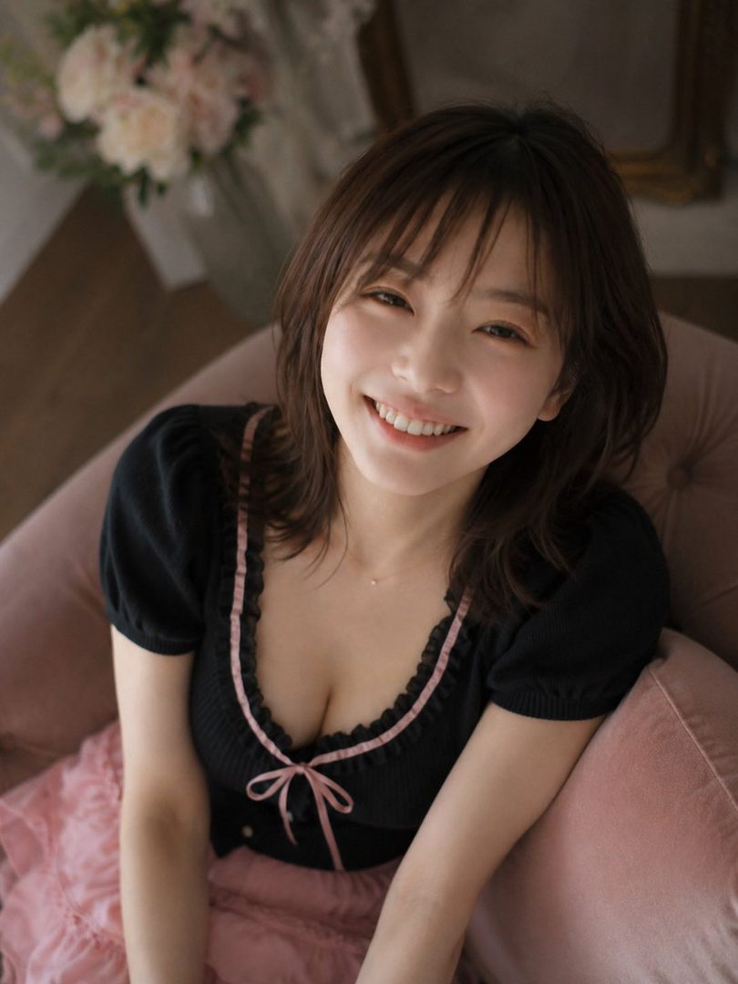 A smiling woman with short brown hair and bangs, wearing a black top with pink trim and a pink skirt, looking up at the camera in a soft-lit indoor setting.