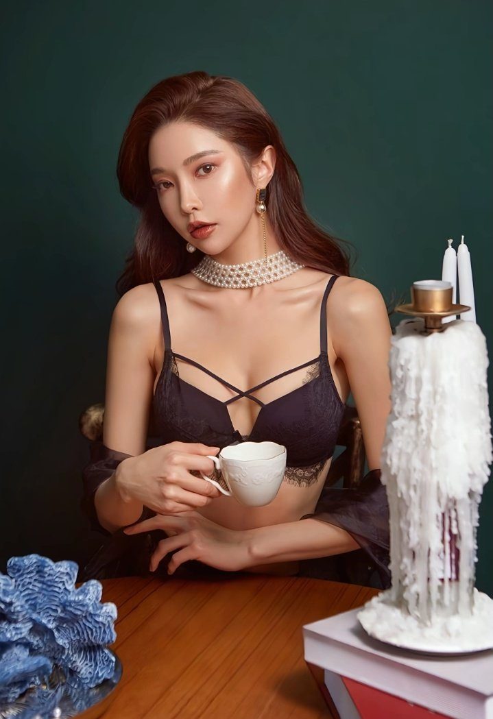 A woman with long brown hair and a pearl choker necklace wears a black lace bra and holds a teacup, sitting at a wooden table with a large candle.