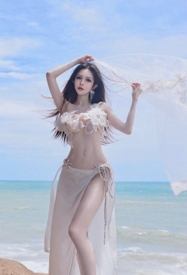 A beautiful woman in a white floral bikini top and sheer sarong poses on a beach with the ocean in the background.