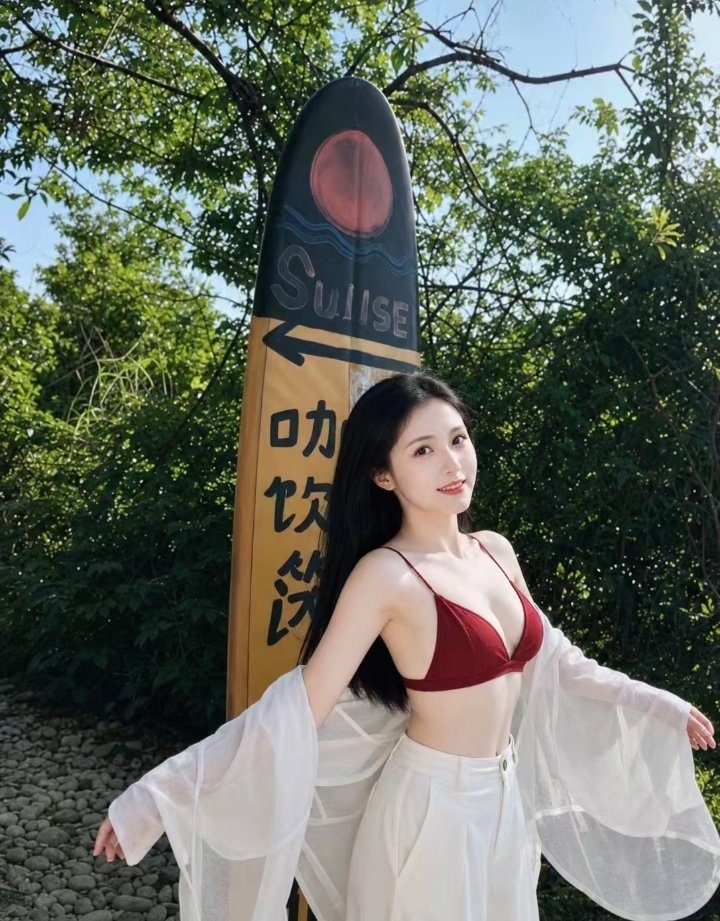 A beautiful woman with long black hair poses outdoors in a red bikini top and white pants, with a surfboard sign behind her.