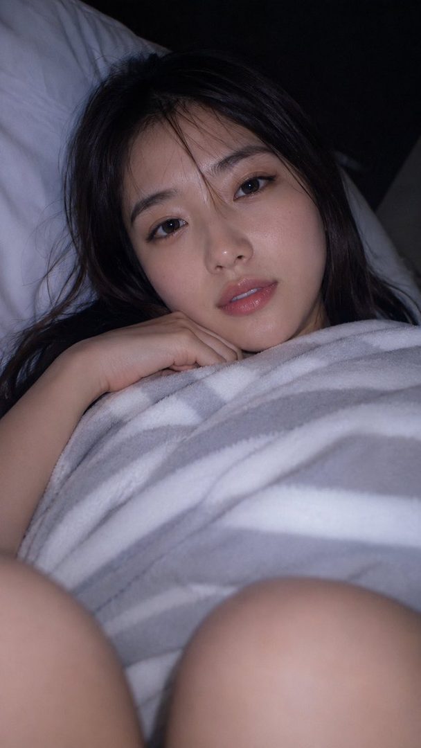 A photorealistic portrait of a young Japanese woman lying in bed, looking calmly at the camera with soft, glossy lips and dark hair, covered by a striped blanket.