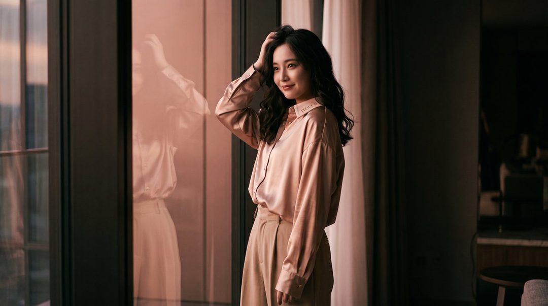 A beautiful Japanese woman with long curly black hair, wearing a champagne-colored satin blouse and beige trousers, stands by a window in a luxurious indoor setting.