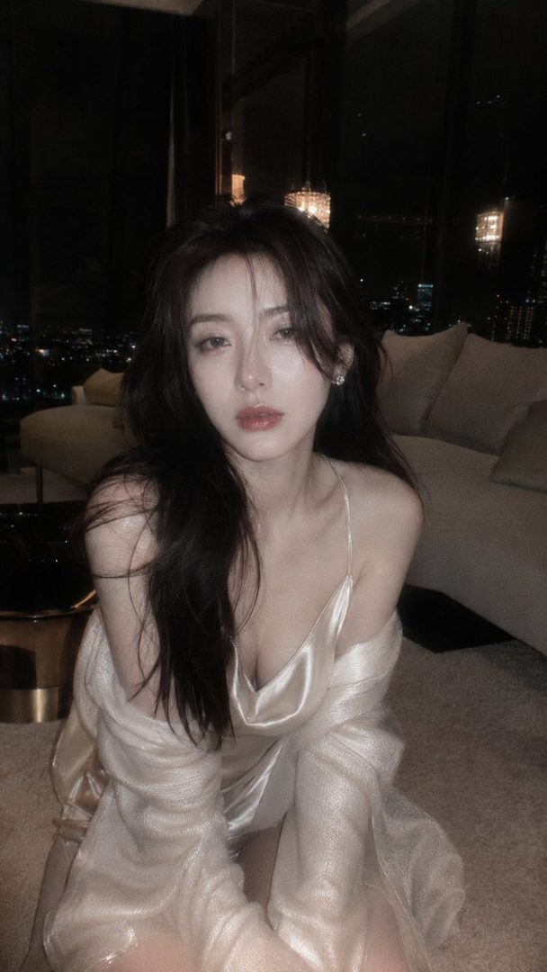 A beautiful East Asian woman with long dark hair wears a satin slip and knit top, sitting on the floor in a luxurious penthouse at night.