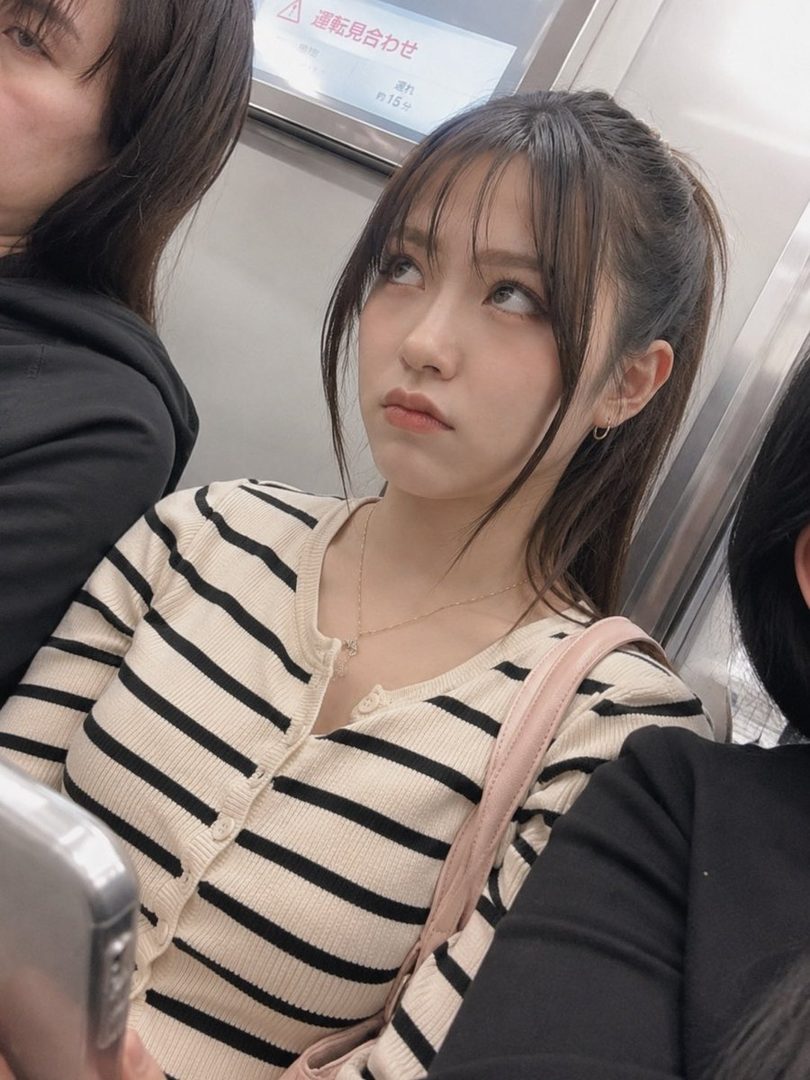 A young woman with dark brown hair in a ponytail and bangs, wearing a striped cardigan, looks up with a pouting expression while on a subway.
