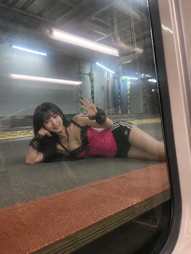 A young woman with long black hair and bangs, wearing a pink corset and black shorts, lies on a subway platform and waves at the camera through a train window.