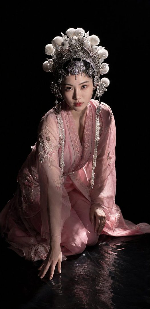 A woman in a pink traditional Chinese dress and an elaborate silver headdress kneels on a dark, reflective surface.