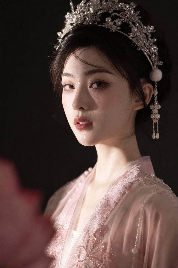 A beautiful woman with dark hair and an elaborate silver headpiece wears a pink traditional Chinese outfit, looking directly at the viewer.