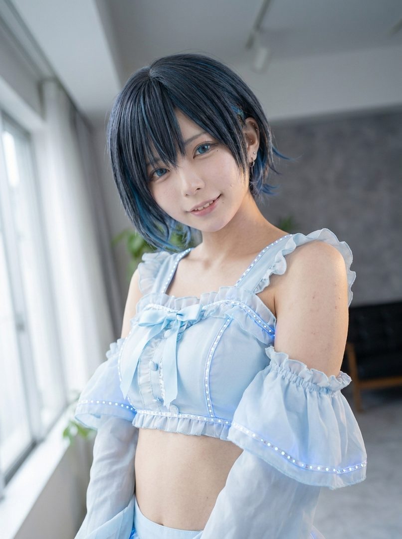 A young woman with short blue hair and blue eyes smiles while wearing a light blue ruffled top with bows and glowing accents indoors.