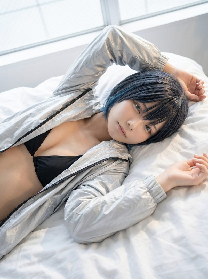 A woman with short black hair and blue highlights lies on a white bed, wearing a black bra and a silver jacket.