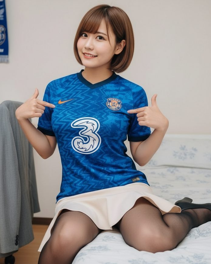 A young woman with short brown hair and bangs smiles while wearing a blue Chelsea FC jersey and a light-colored skirt, sitting indoors.