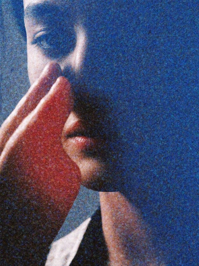 A close-up portrait of a person with their hand near their face, rendered with a grainy, cinematic effect and deep blue and red tones.