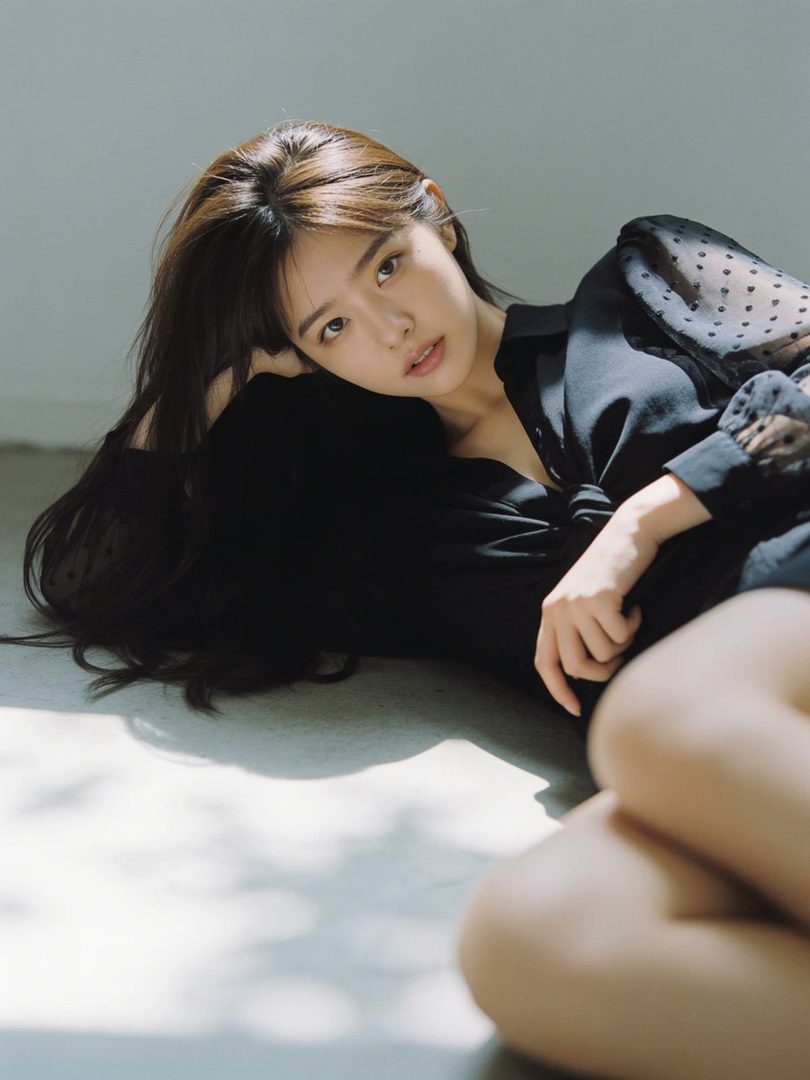 A beautiful East Asian woman with long brown hair is lying on the floor, wearing a black sheer blouse, with soft light and shadows on the floor.