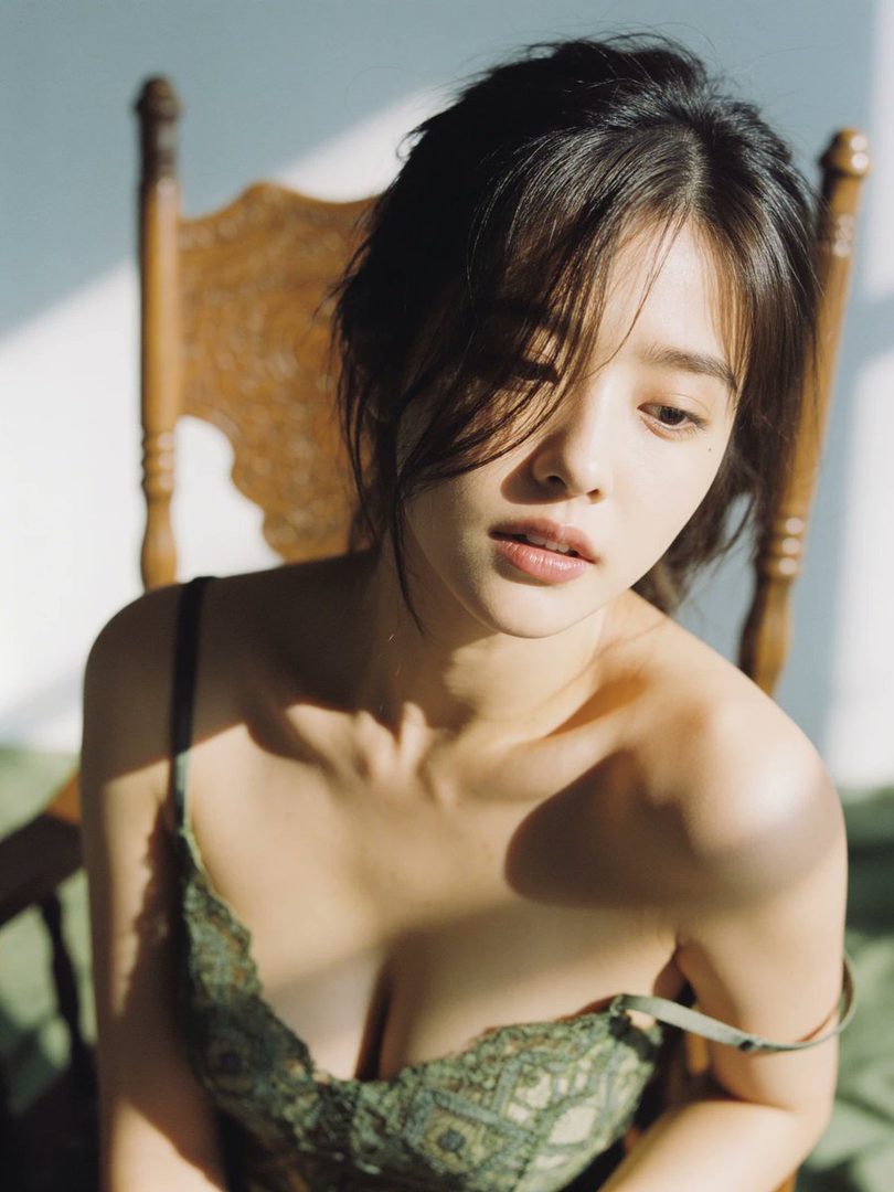 A beautiful woman with brown hair sits indoors, bathed in natural light, wearing a green lace top.