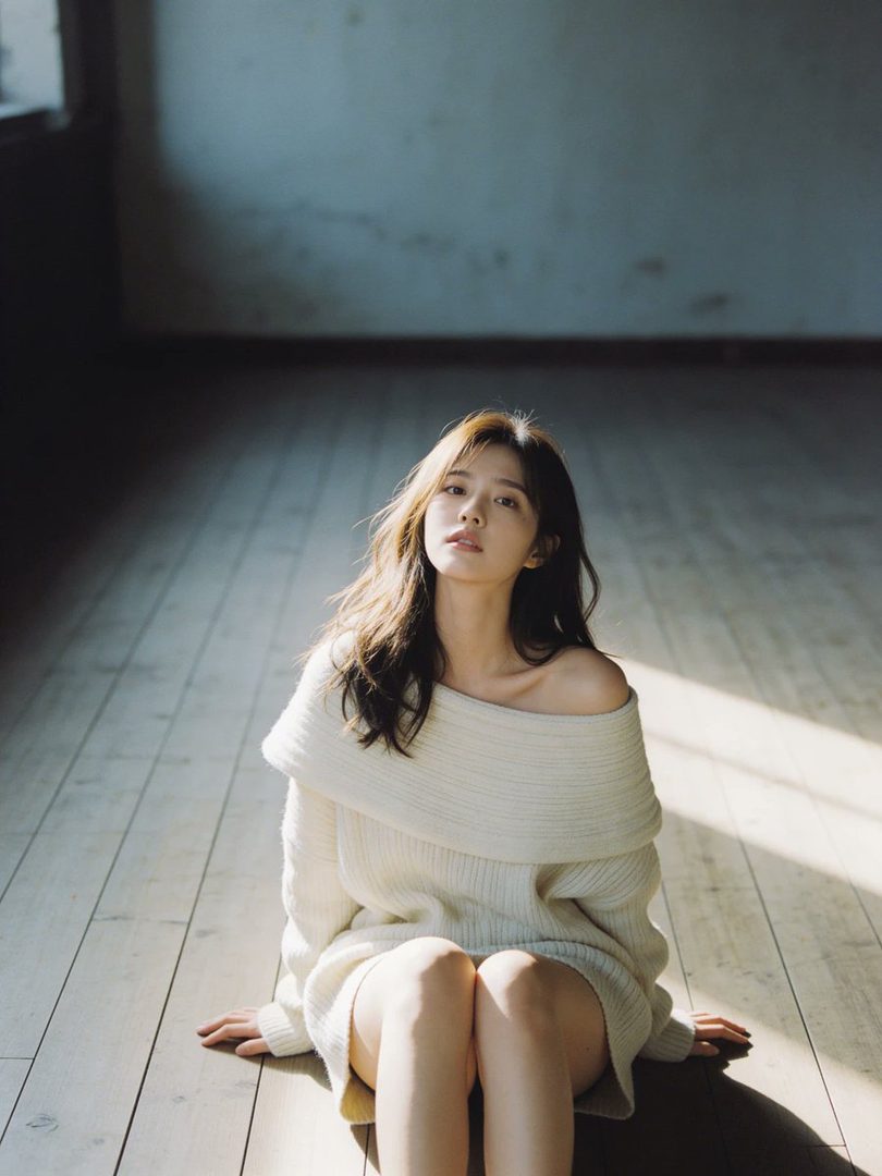 A young woman with long brown hair sits on a wooden floor in a sunlit room, wearing an off-the-shoulder white sweater.