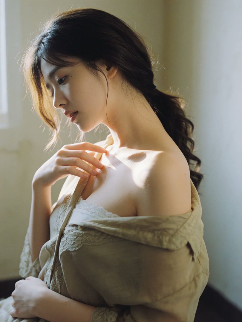 A beautiful woman with long brown hair in a ponytail, wearing a light-colored top, is bathed in soft morning light indoors.