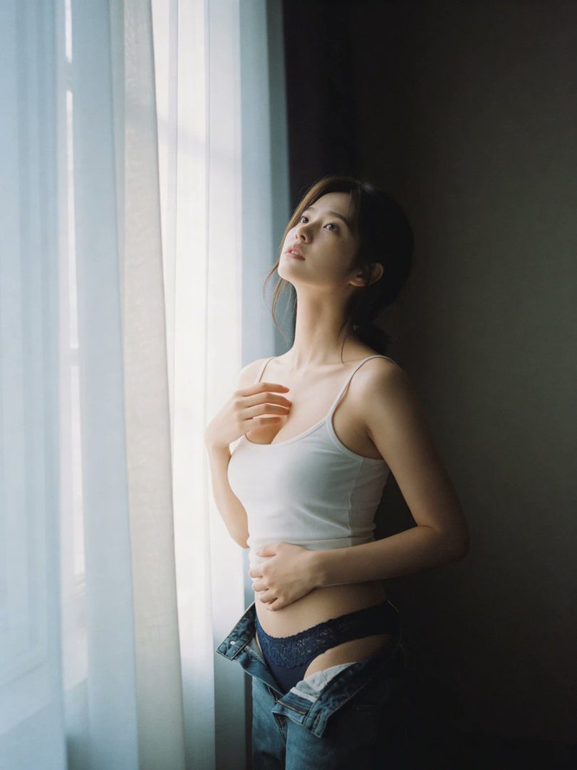 A woman in a white camisole and unbuttoned jeans stands by a window, looking up with soft light on her face.
