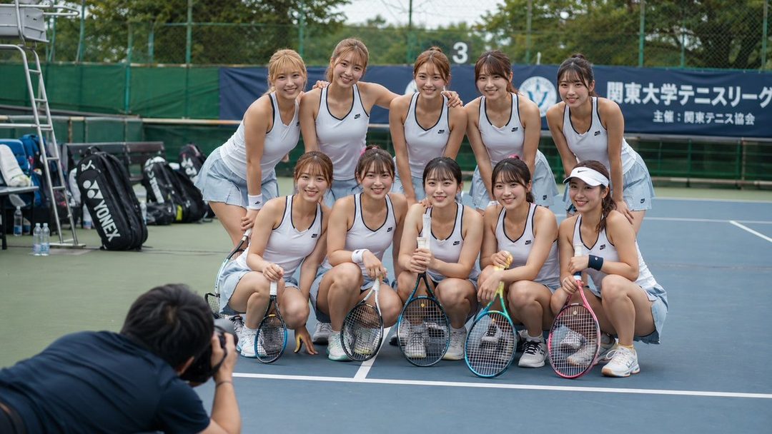 A group of ten young women in tennis outfits pose for a photo on a tennis court, holding rackets and smiling.