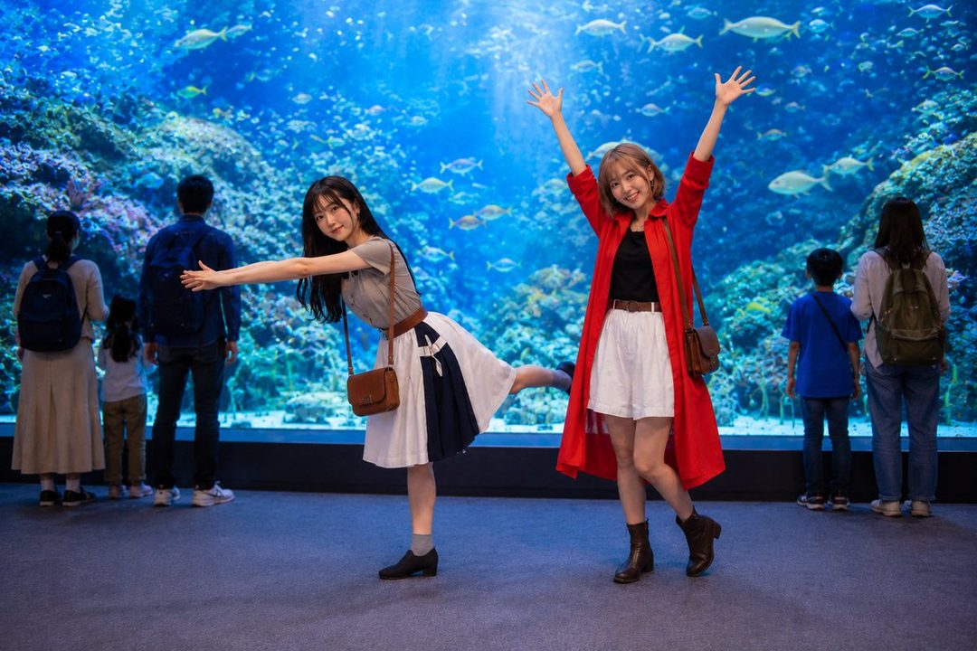 Two young women pose playfully in front of a large aquarium tank filled with fish and coral.