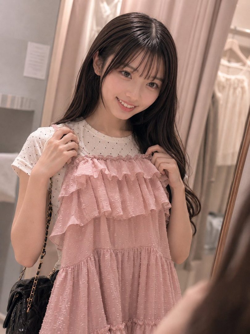 A young woman with long brown hair and bangs smiles while holding up a pink ruffled dress in what appears to be a fitting room.