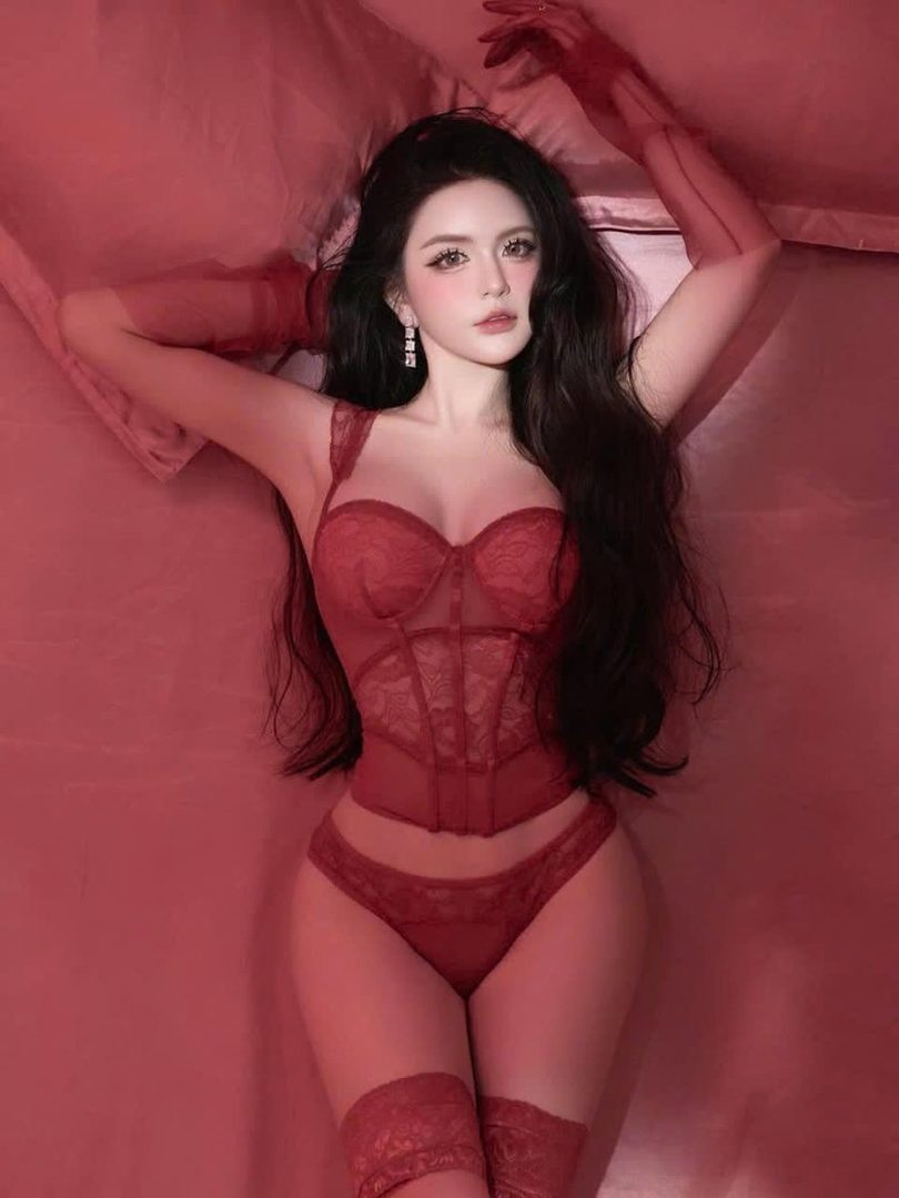 A woman with long black hair is lying on a red bed, wearing red lingerie and thigh-high stockings.