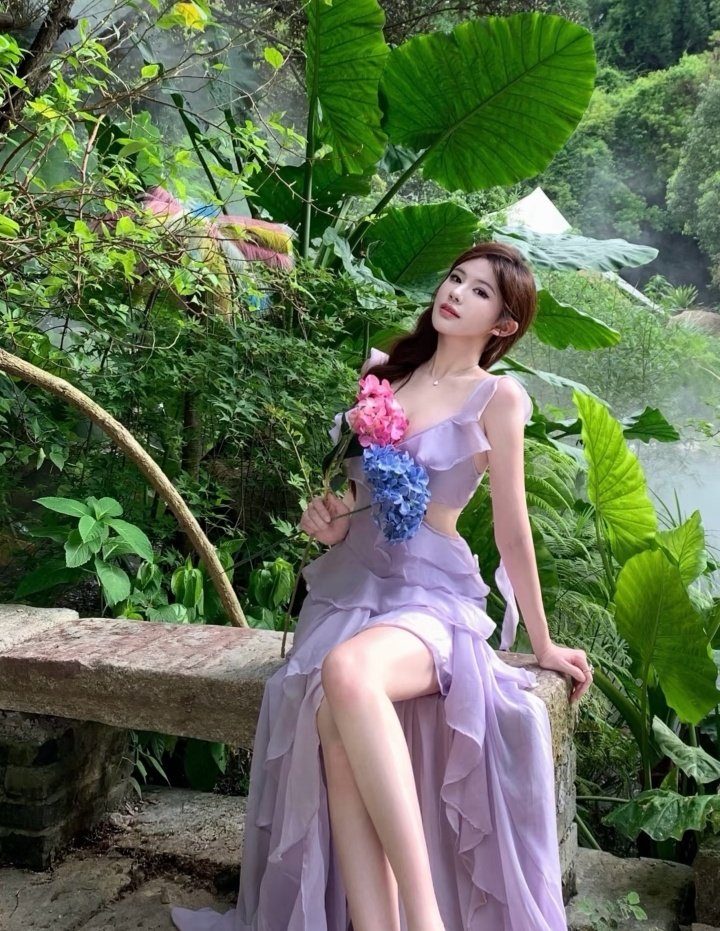 A woman in a flowing lavender dress sits on a stone bench amidst lush green foliage, holding pink and blue flowers.