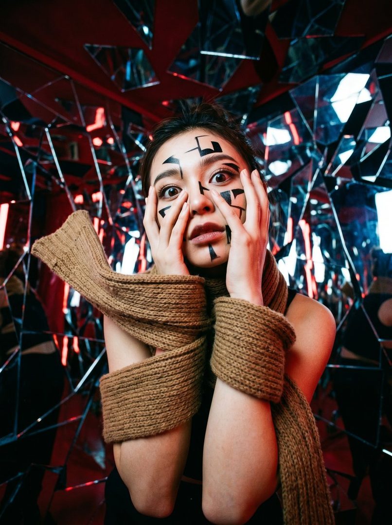 A woman with black markings on her face and a brown knitted scarf wrapped around her arms, looking surprised in a room with mirrored walls.