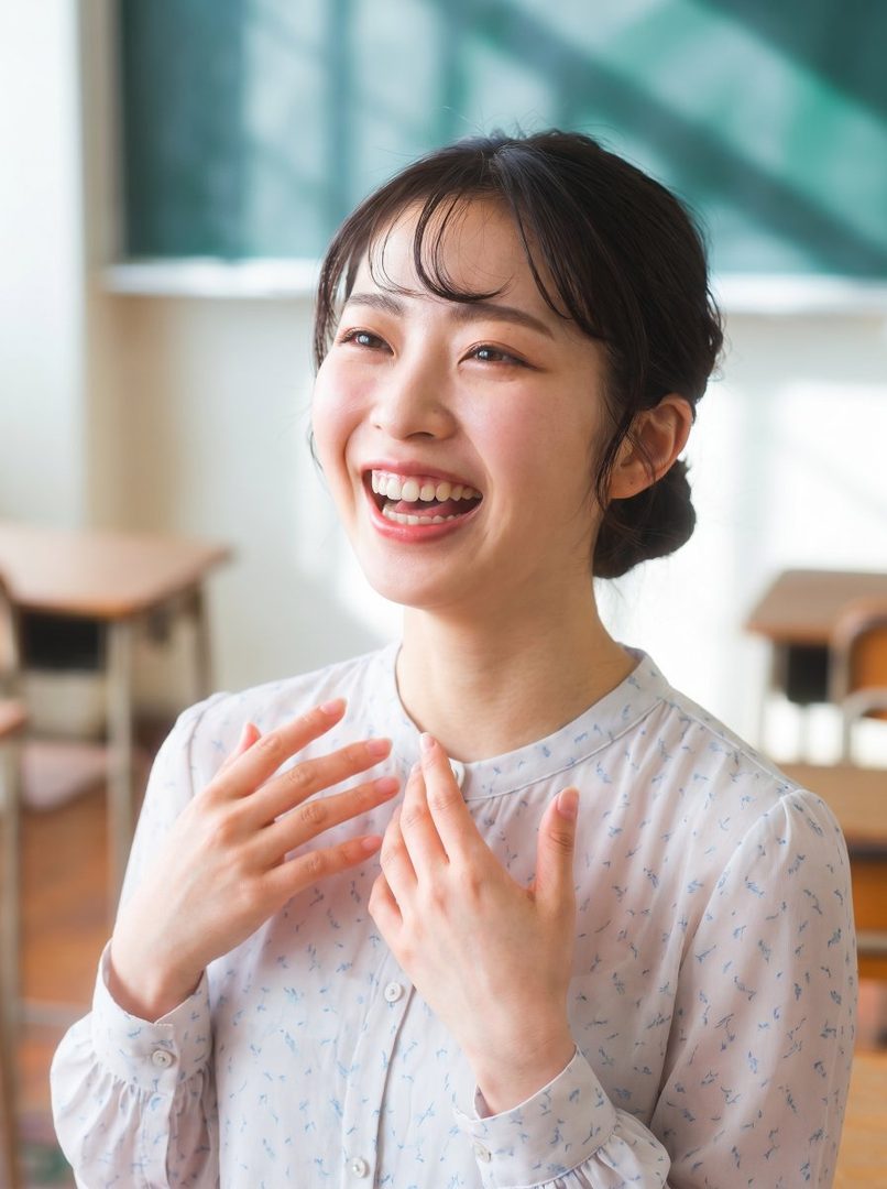 A smiling young East Asian woman in a white patterned blouse, with her hands near her chest, in what appears to be a classroom setting.