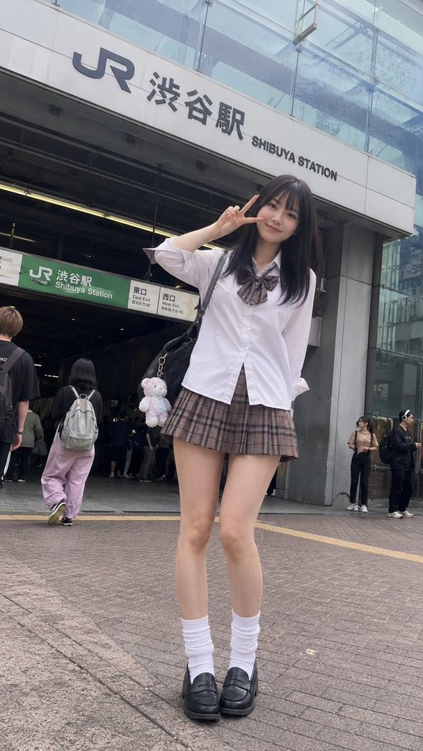 A young woman in a Japanese school uniform poses with a peace sign in front of Shibuya Station.