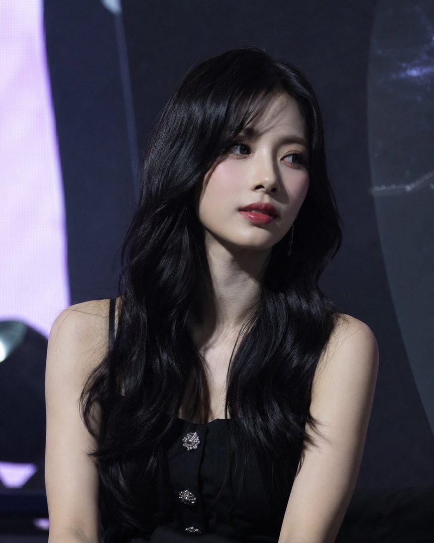 A beautiful East Asian woman with long black hair and a black dress looks to the side in a dimly lit setting.