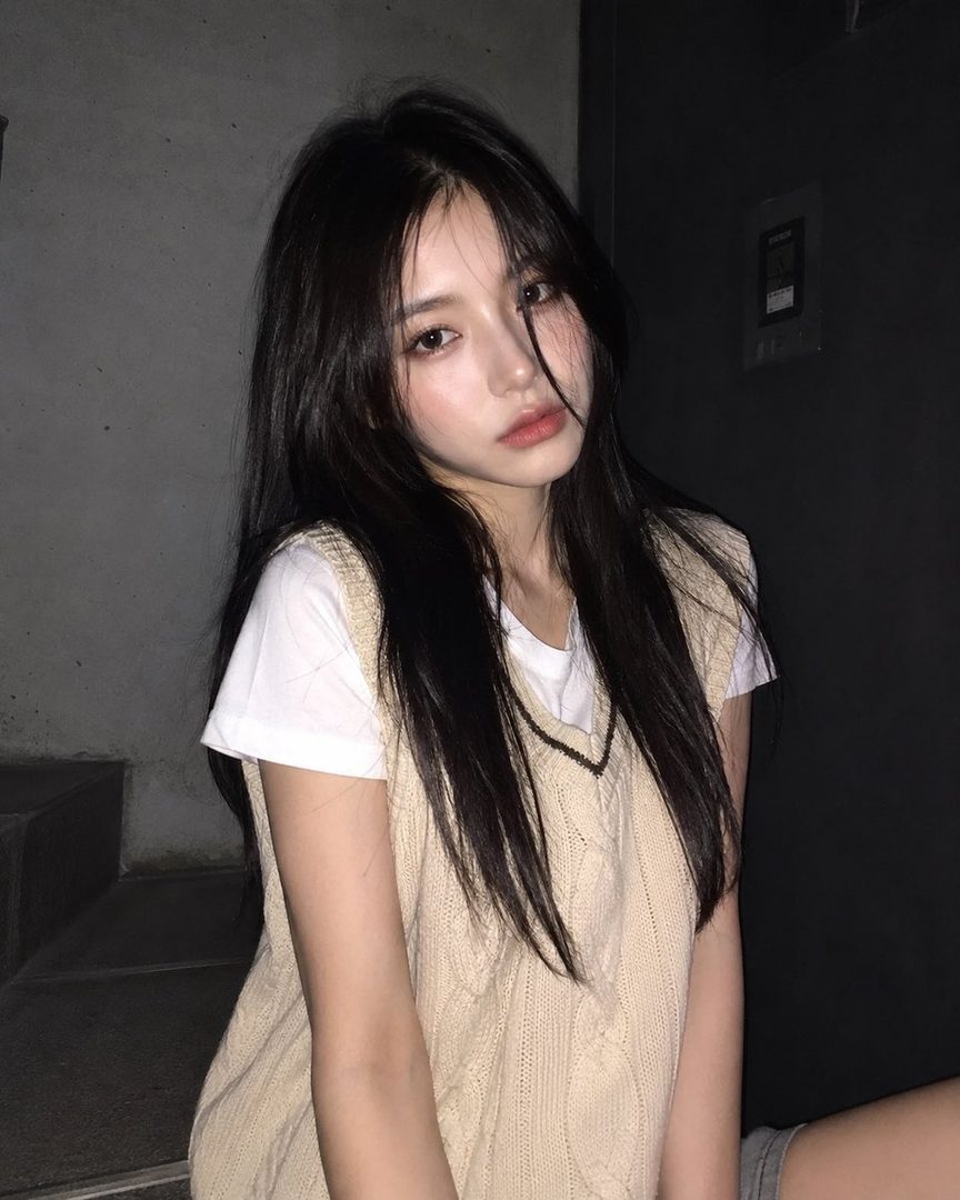 A photorealistic AI portrait of a young woman with long black hair, wearing a white t-shirt and a beige knit vest, looking directly at the viewer.