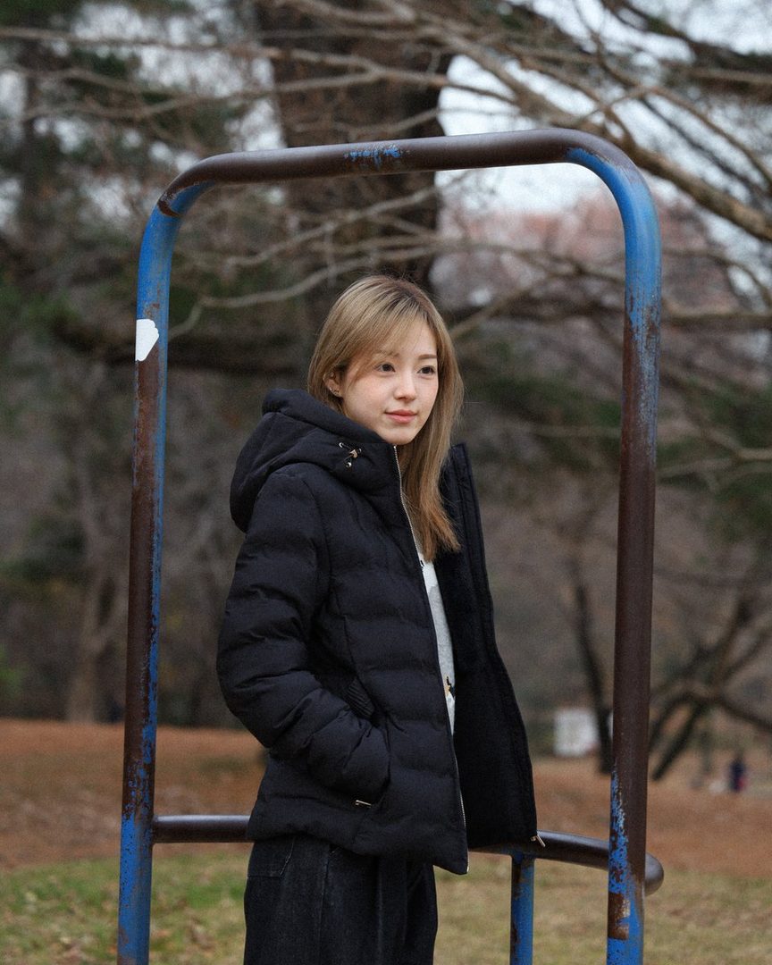 A young woman with blonde hair wearing a black puffer coat stands outdoors in a park.