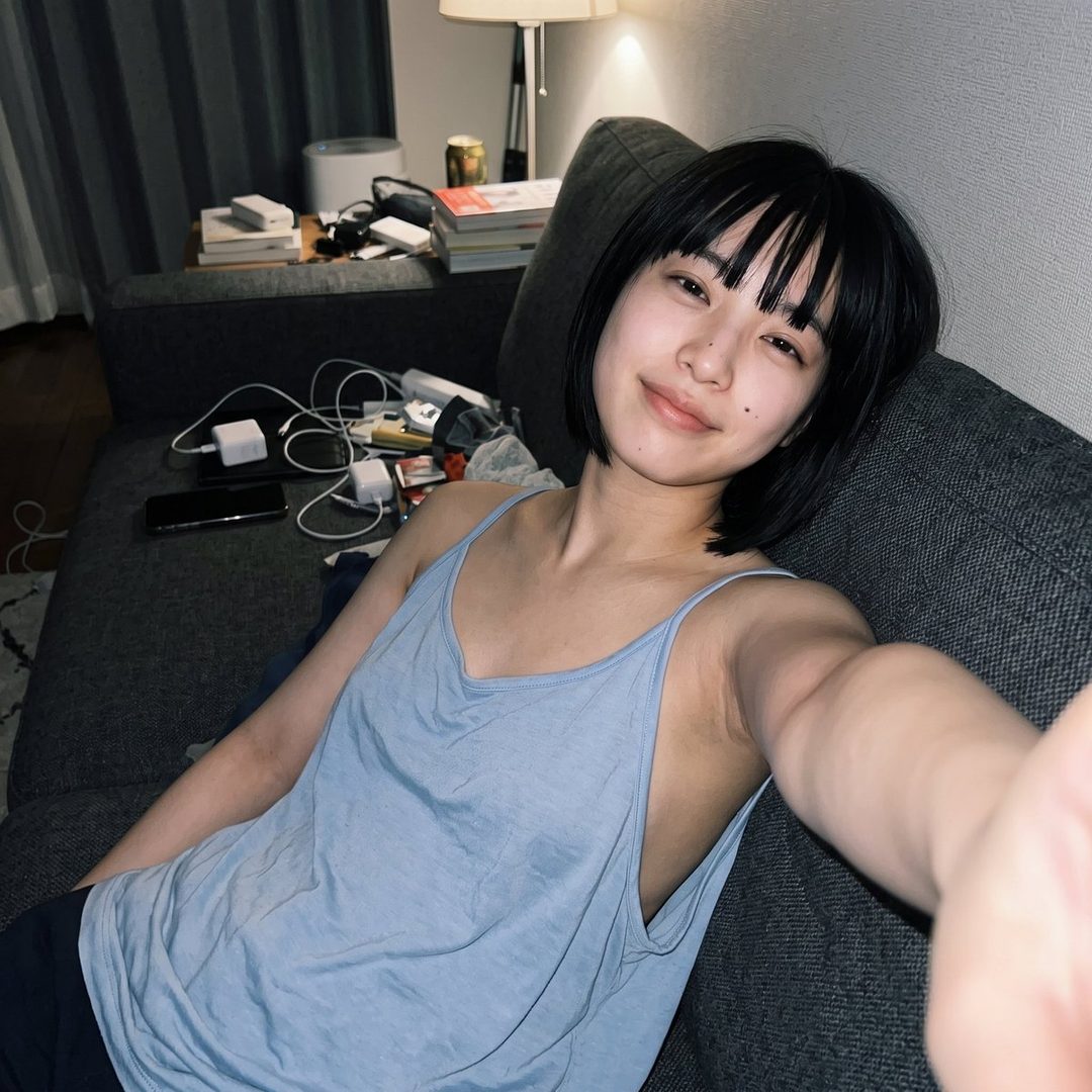 A young woman with short black hair and bangs takes a selfie while relaxing on a sofa indoors.