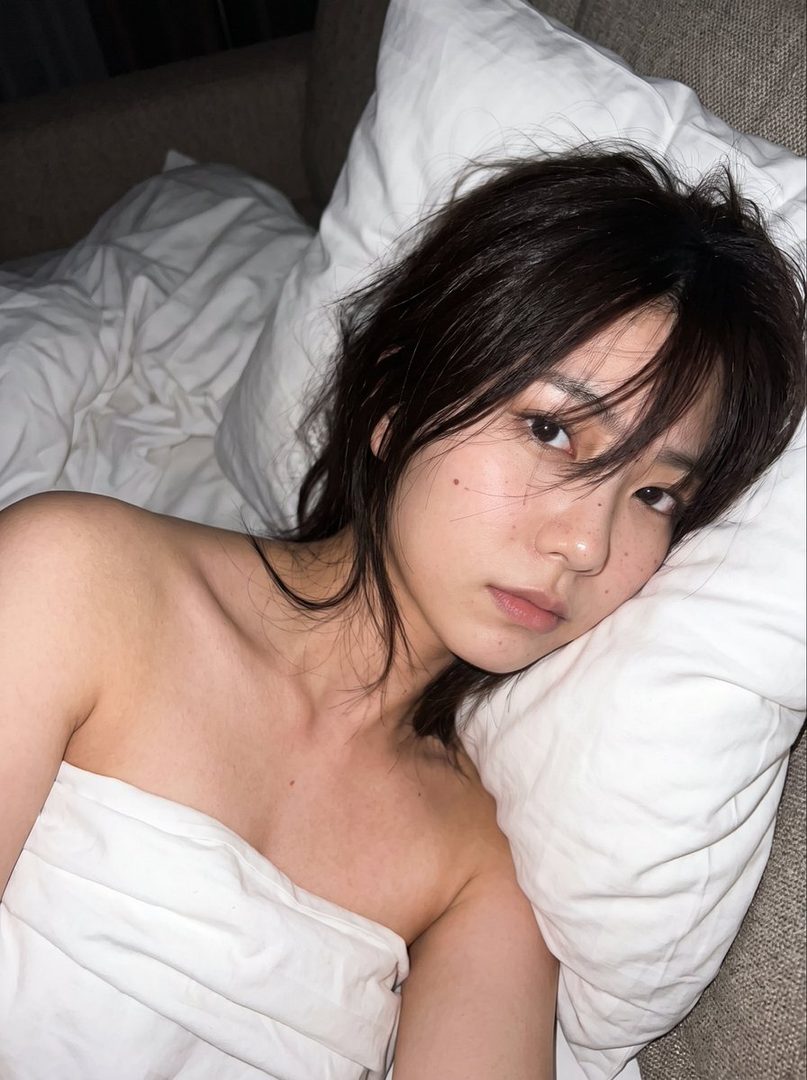 A photorealistic portrait of a young woman with brown hair lying in bed, covered by a white sheet.